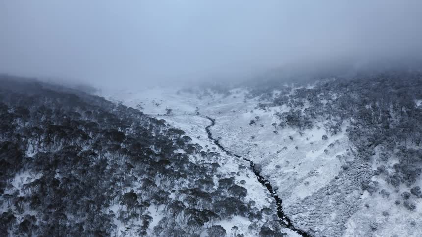 Aerial view of a river flowing at the bottom of a small valley between two mountains covered in snow under the clouds
