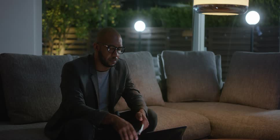 Man with shaved head glasses beard and green blazer jacket sits on gray couch using smartphone and tablet in living room at home