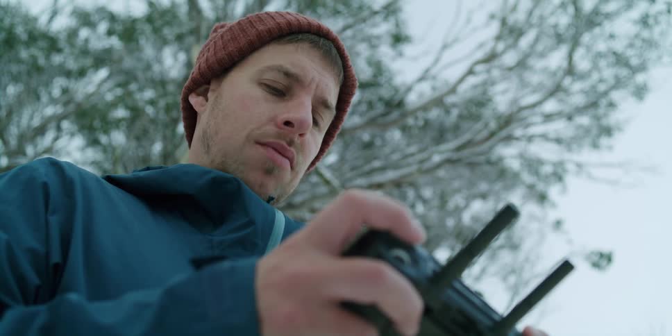 Young man wearing winter clothes stands under a tree covered in snow controlling a drone with a remote