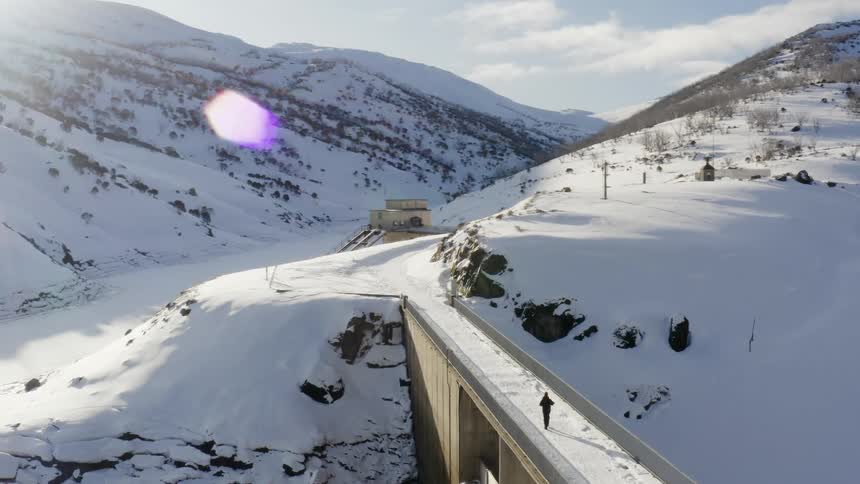 Orbiting aerial view of a man crossing a bridge to a remote station on the snow covered slopes of a mountain at morning