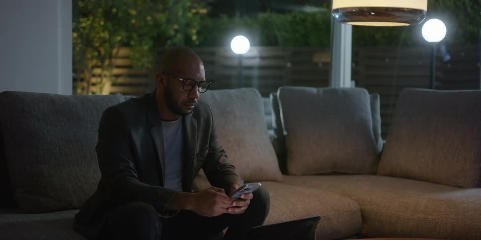 Man with shaved head glasses beard and brown blazer jacket sits on gray couch using smartphone in living room at home