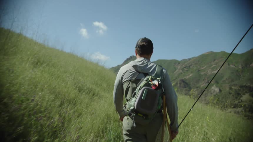 Young man carrying fishing equipment walks on the slopes of a mountain at daytime