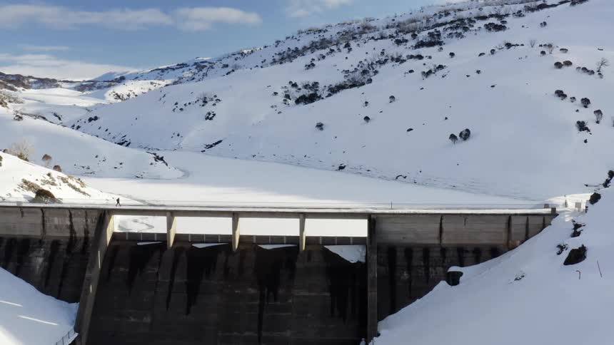 A person is walking across a bridge above a dam on a snowy winter day and there is a mountain in the distance