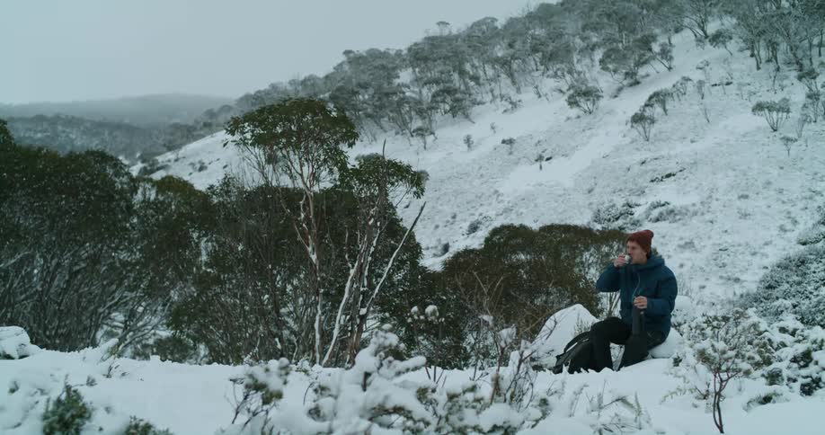 A man drinks from the cap of an insulated bottle while sitting on a snowy mountainside during the day
