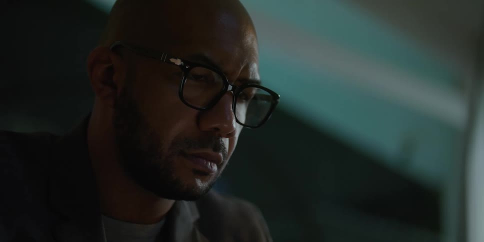 Man with shaved head glasses beard and blazer jacket looks down at laptop screen in living room at home