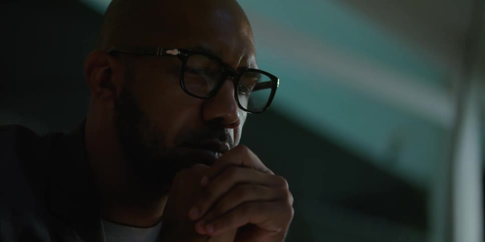 Man with shaved head and glasses and beard looks down at laptop screen in living room at home