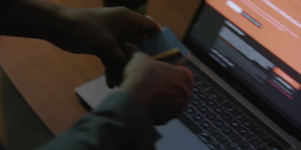 A man sitting in front of a laptop picks up a small notebook and a pencil and puts it in his pocket