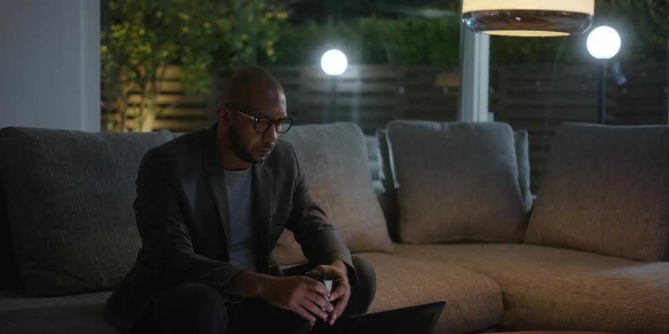 Bearded bald man wearing glasses enjoys a drink while sitting on a couch at home at night