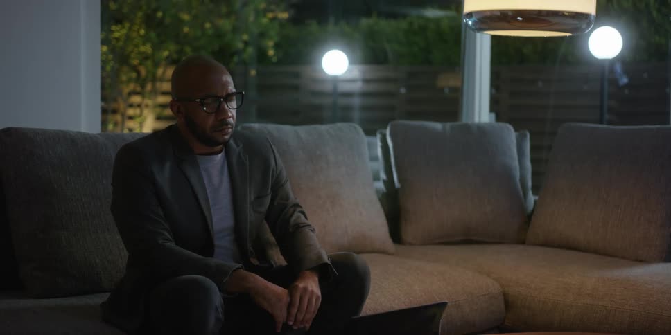 A man typing on a laptop while sitting on a living room sofa at night