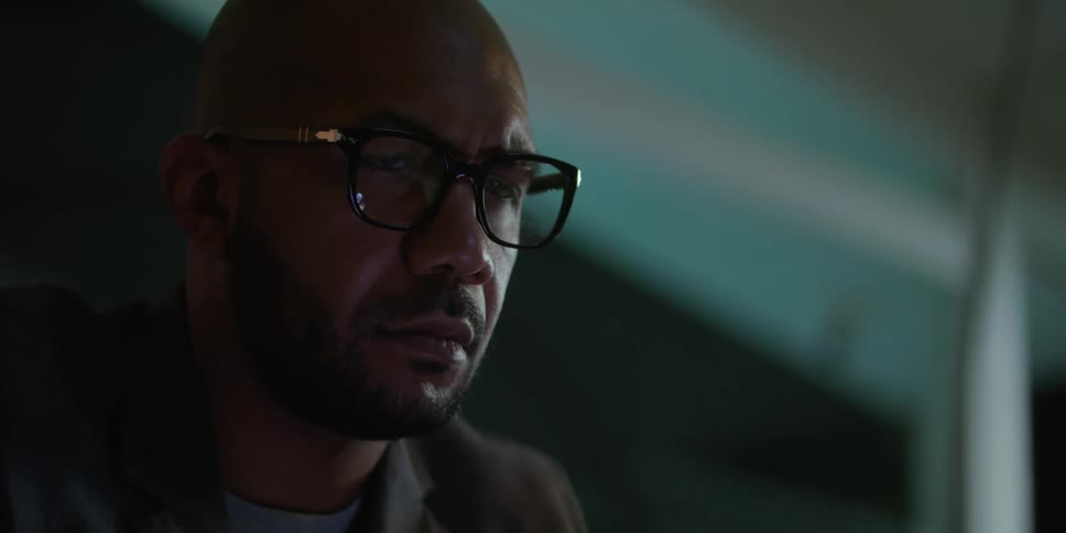 Close up of the face of a bald bearded man wearing glasses sitting on the living room at night