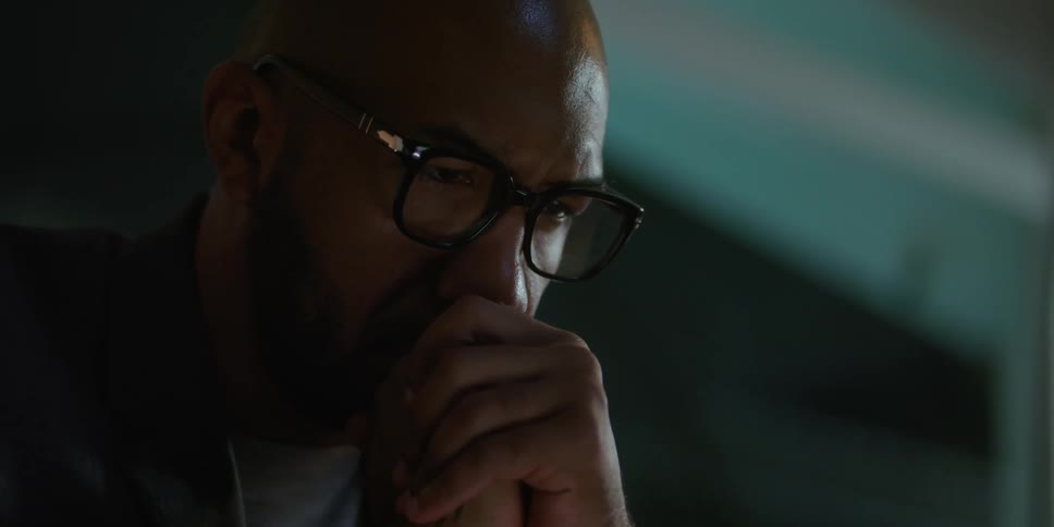Man with shaved head glasses beard and brown blazer jacket looks down and puts his hands on his mouth in room