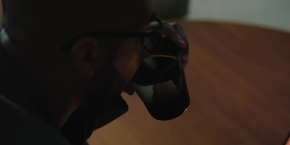 A man sets down his cell phone and clasps his hands and looks at his laptop then picks up a glass of water and takes a drink