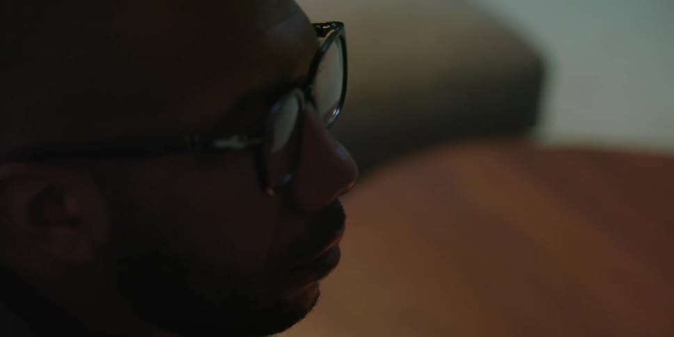 A man with glasses sits at a table thinking and then gets up from his chair and walks away