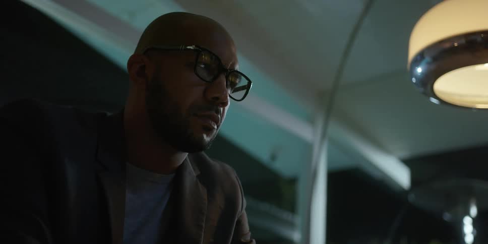 Bearded bald man wearing glasses checks his phone while sitting on a couch at home at night