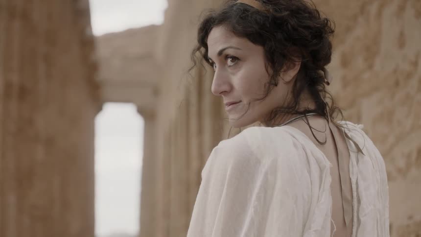 A woman looks over her shoulder while standing near ruins