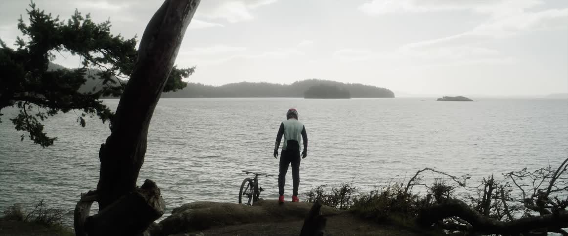 Cyclist stands on shore next to bicycle looking out to water across from misty promontory
