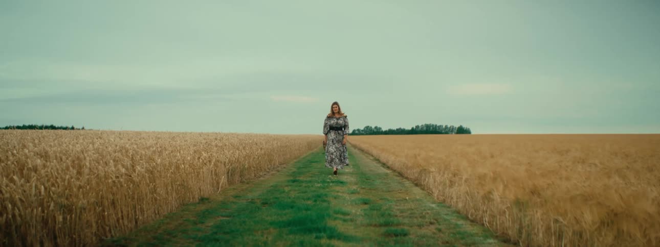 Woman moves along grass aisle between crop fields under an overcast sky