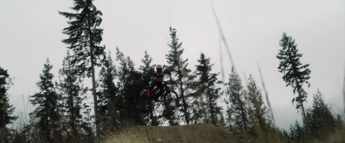 A cyclist does a trick in the air after going airborne while writing a bike up a dirt ramp in a woodland on a cloudy day
