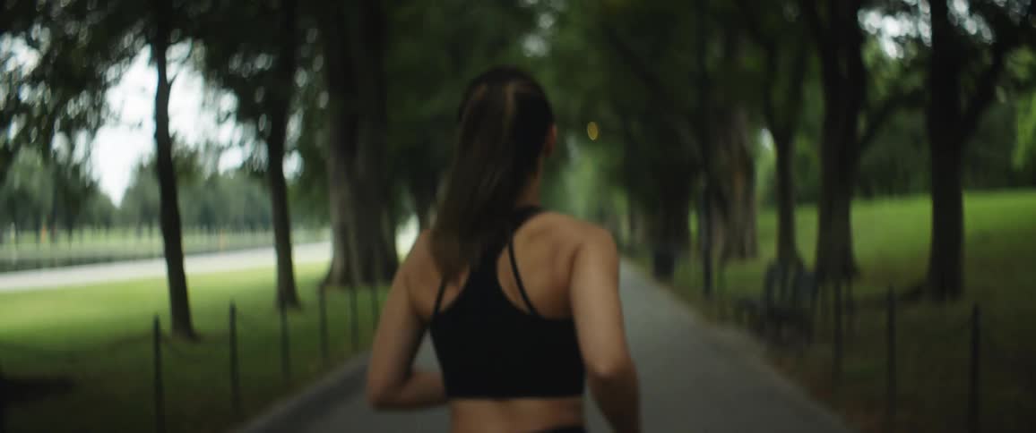 A woman is standing on a walkway between trees in a park and she turns and jogs away