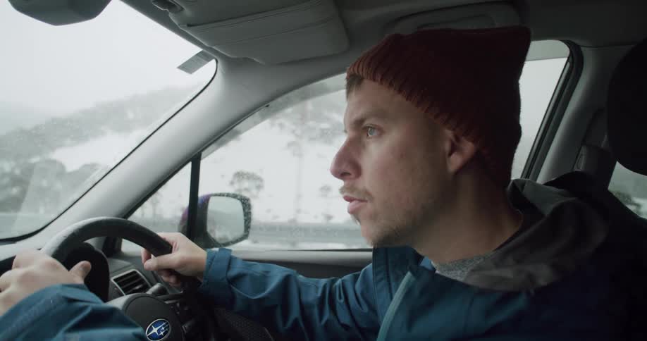 A man is driving an automobile on a road near snow covered mountains