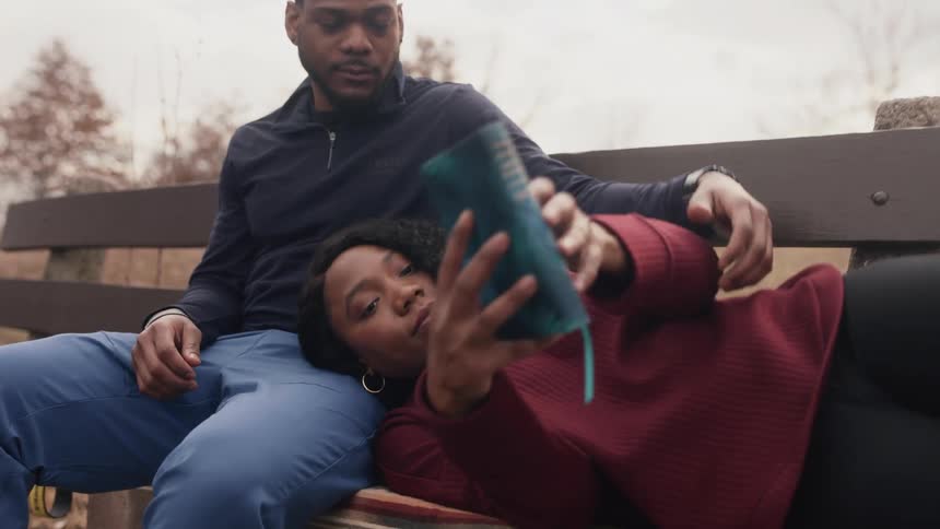 A woman rests her head in the lap of a man sitting on a bench outdoors as she reads a book