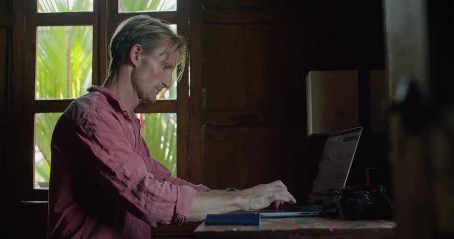 Man with pink shirt sitting at desk types on laptop in room