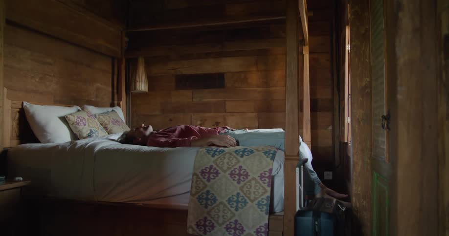 Tired man on vacation sitting on a bed on a small bedroom lays down to rest