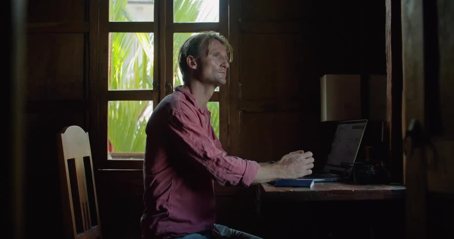 A man is sitting at a table typing on a laptop computer and he folds his hands as he looks at the screen
