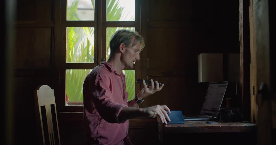 A man is sitting at a table typing on a laptop computer and he picks up his phone and reads from a small book on his desk