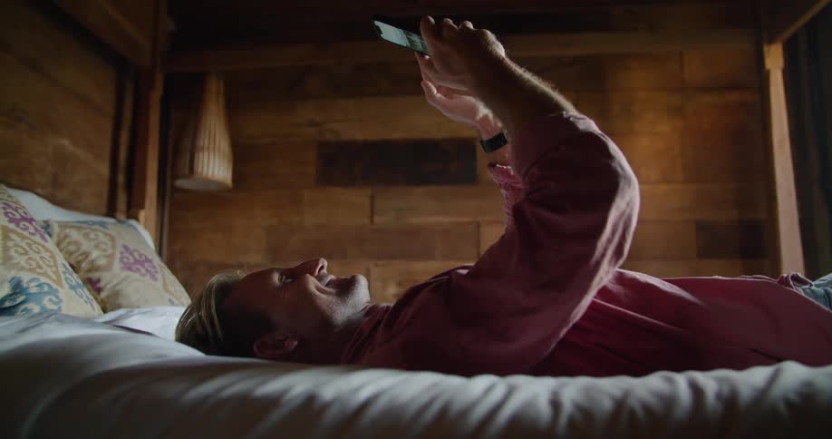 A smiling man scrolls through his smartphone while lying on a bed during the day