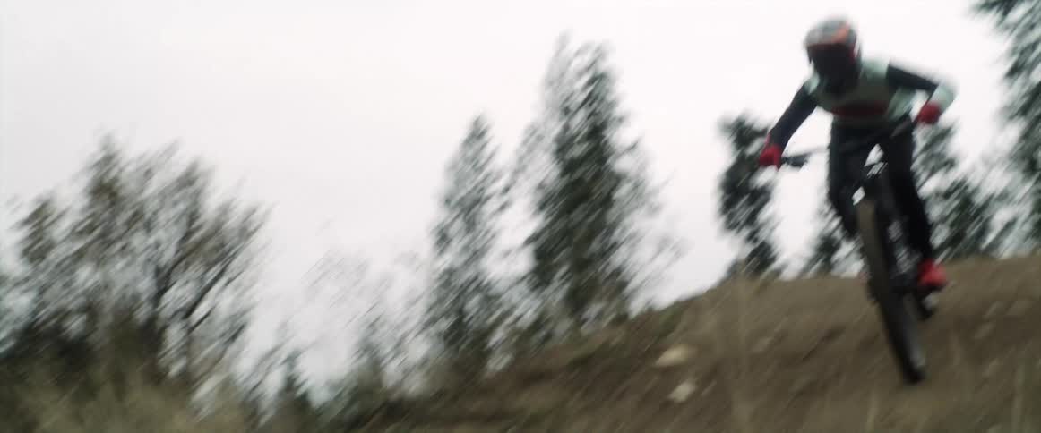 A cyclist rides on a trail and flies over a dirt ramp in the woods and turns in mid air