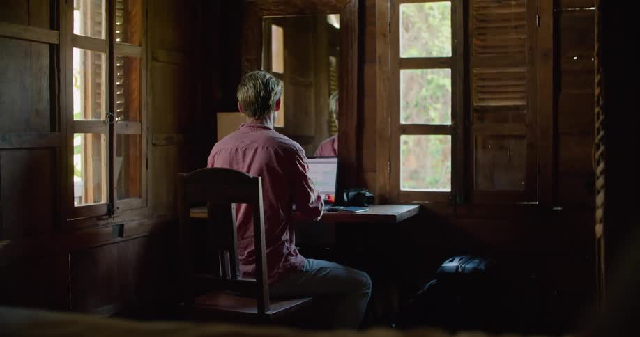 A man sitting at a desk at home leans on his hand as he thinks then types on a laptop