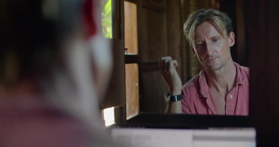 A man is sitting at a table typing on a laptop computer and his reflection is in the mirror in front of him