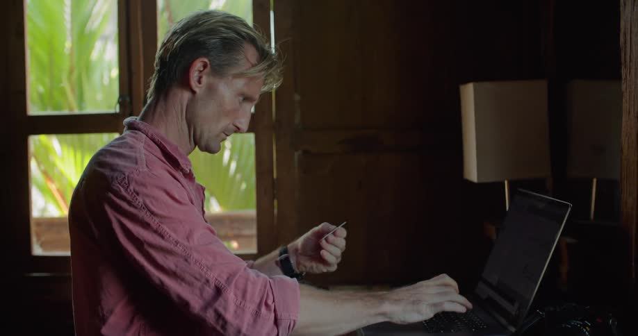 Man with pink shirt and wristwatch takes credit card out of his pocket and enters information on laptop