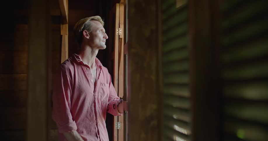 A man pushes his window open then smiles as he sits on the sill