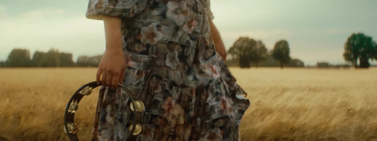 Woman wearing a long dress walks near a wheat field with a tambourine on her hand