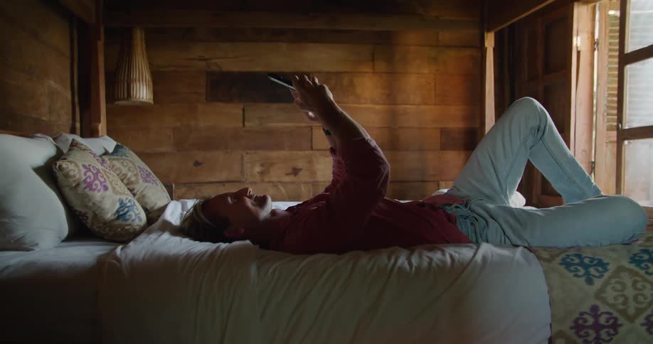 Man on vacation resting on the bed on a hotel bedroom comfortably texting on his phone