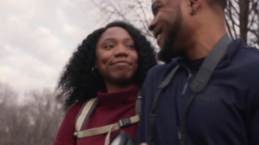 Man with beard black top and camera takes a walk with woman with curly hair and red turtleneck sweater in the forest