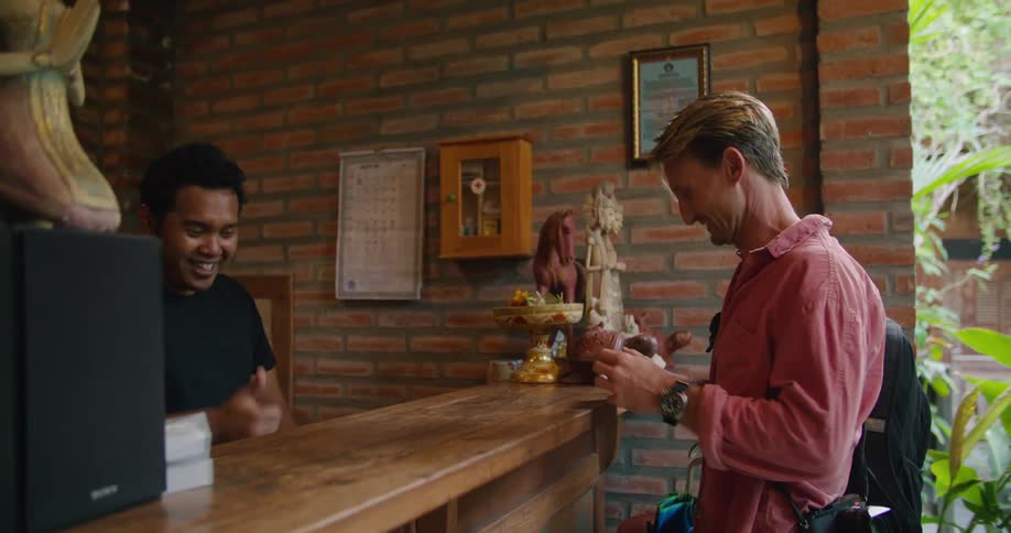 A man with a backpack on his should walks up to a counter and shakes hands with the man there and talks to him as he places his credit card on the counter