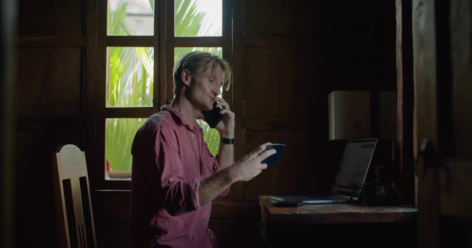 A man is sitting at a table with a laptop computer and talking on his phone as he looks at a small book