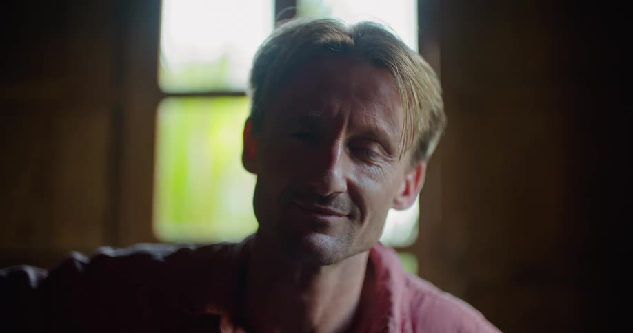 Blonde man with pink shirt smiles at camera in front of windows in room