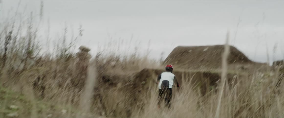 Person rides dirt bicycle along narrow dirt path and up mound to perform a jump