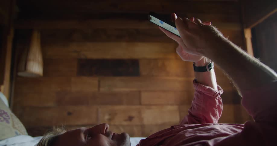 Man on vacation resting on the bed on a hotel bedroom comfortably browsing on his phone