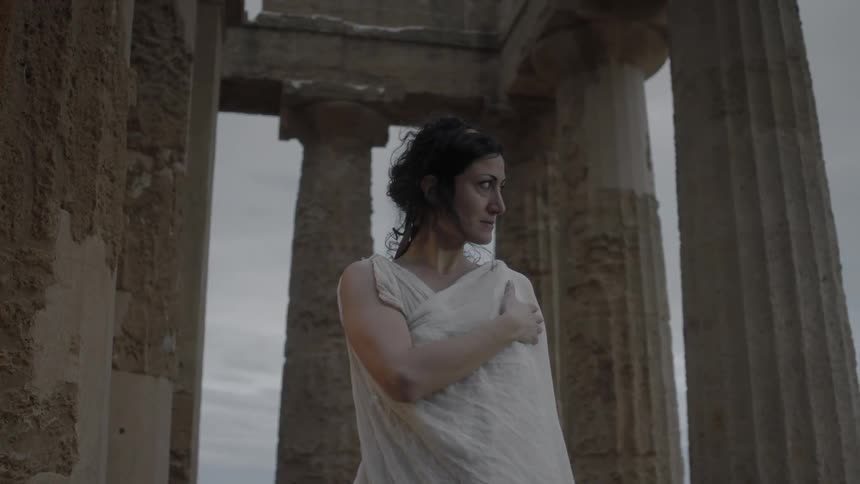 Woman in a white toga stands in the middle of an old structure with columns
