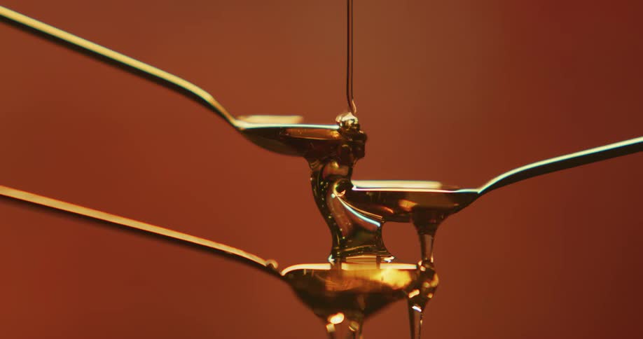 Thick amber honey dripping down over stacked spoons in front of a brown backdrop