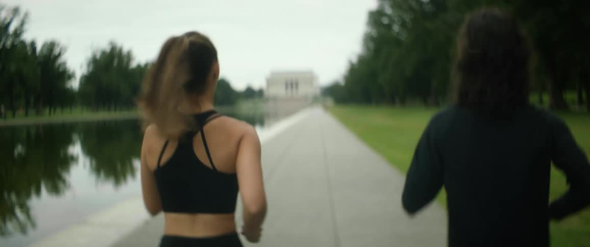 Woman with ponytail black sports bra and man with long wavy hair and black sweater jogs by lincoln memorial reflecting pool