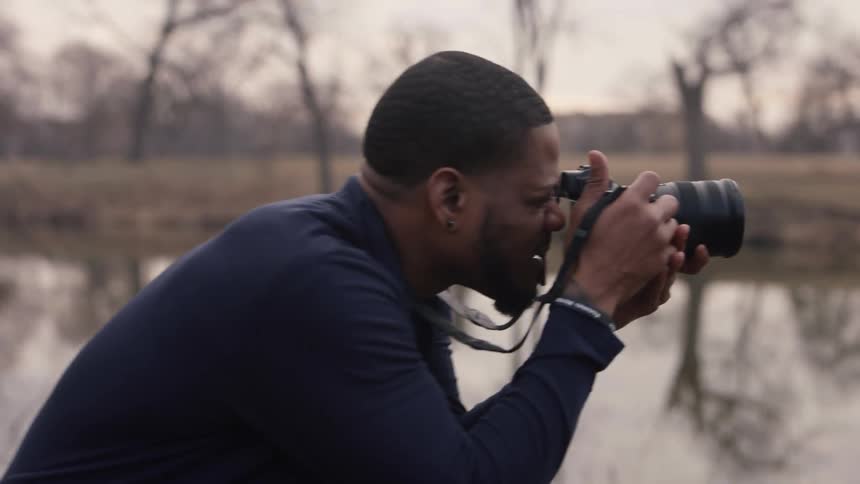A photographer raises a camera to take photographs on the shore of a pond
