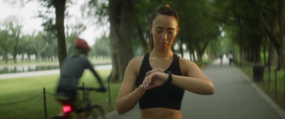 A woman is standing on a sidewalk in a park looking at her watch as people pass by her