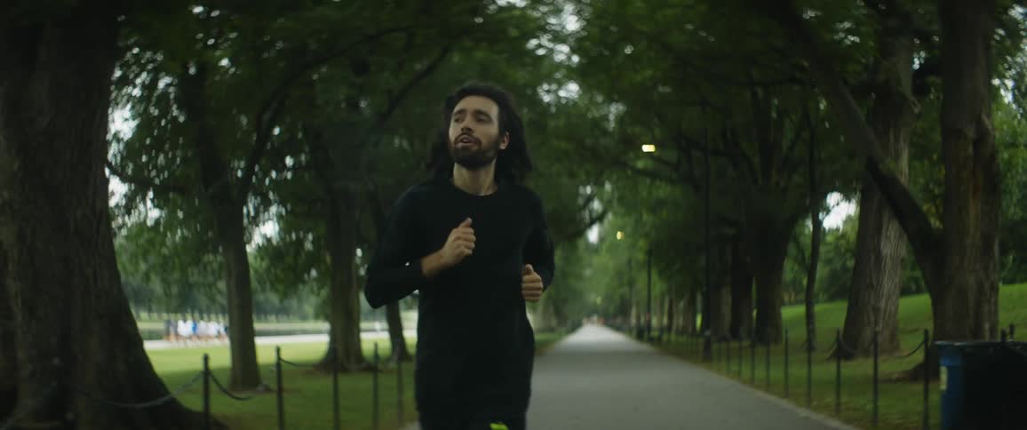 A man is jogging on a walkway between trees in a park as a woman passes by