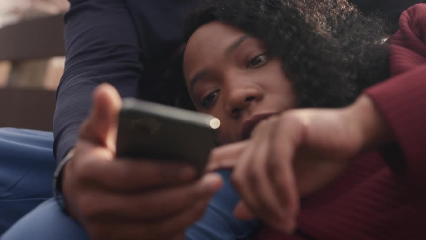 Woman with curly hair and redturtleneck sweater puts her head on mans lap with jeans and black shirt sitting on bench using smartphone